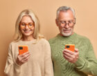 Elderly spouses use modern technologies. Old woman and man hold mobile phones download new application browse socail networks wear spectacles and casual jumper stand indoor. Generation and tech