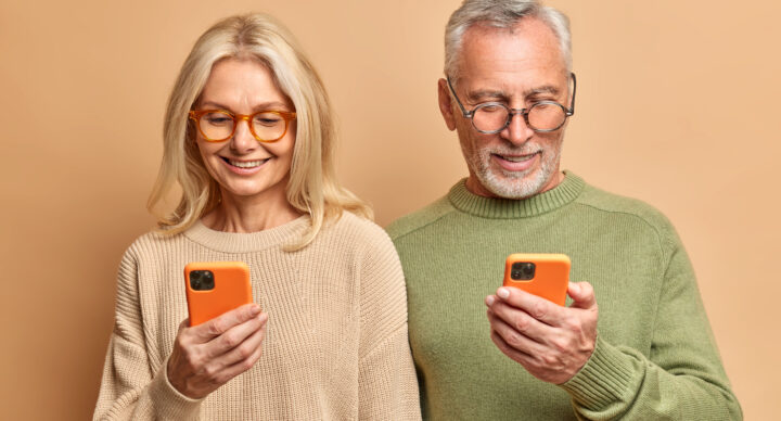 Elderly spouses use modern technologies. Old woman and man hold mobile phones download new application browse socail networks wear spectacles and casual jumper stand indoor. Generation and tech